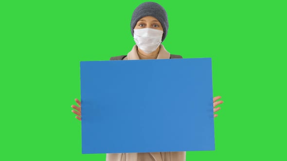 Woman in Outdoor Clothes and in Medical Mask Holding Blank Board on a Green Screen, Chroma Key.