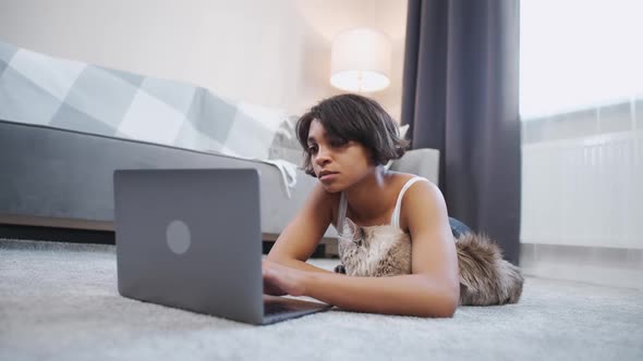 Young Darkskinned Woman Lies on the Floor in the Living Room with a Cat and Works at a Laptop