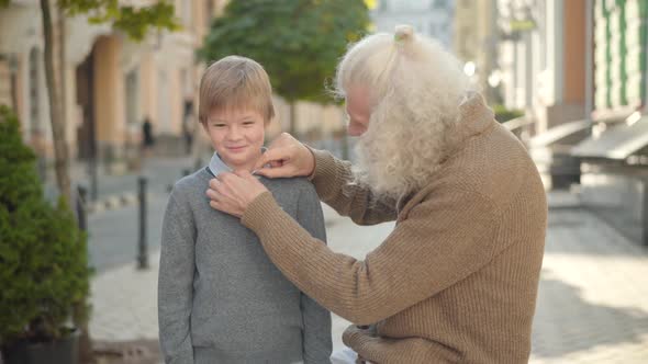 Caring Old Caucasian Man Adjusting Shirt of Smiling Little Boy on City Street. Thankful Grandson