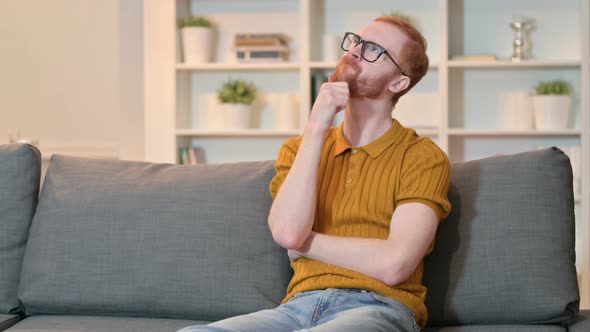 Pensive Redhead Man Sitting on Sofa at Home and Thinking 