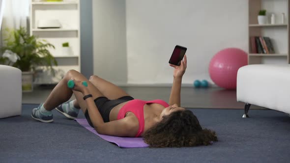 Woman Lying on Floor, Lifting Dumbbell with One Hand and Using Mobile Phone