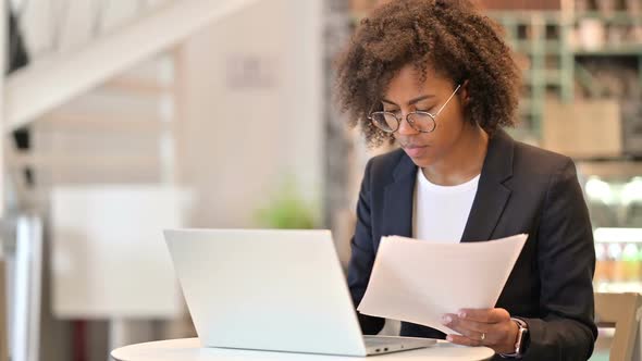 Focused African Businesswoman with Laptop Reading Documents in Cafe