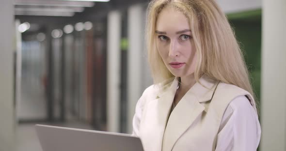 Portrait of Beautiful Blond Caucasian Woman Standing with Laptop in Open Space Office, Looking at