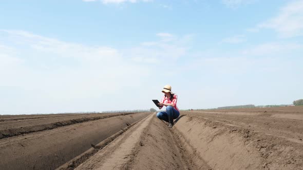 Female Farmer, Agronomist Sits Between Special Soil Rows on Field. She Tests, Using Tablet, Quality