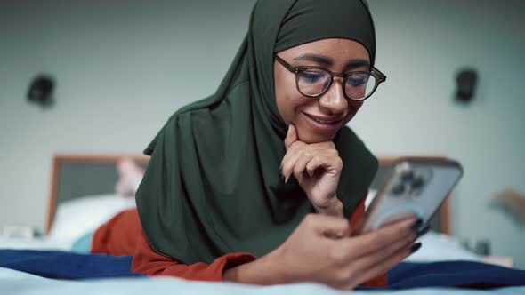 Positive Muslim woman wearing eyeglasses typing on phone