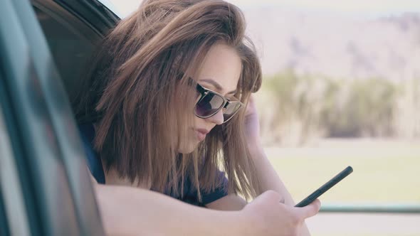 young woman uses mobile phone leaning out of car window