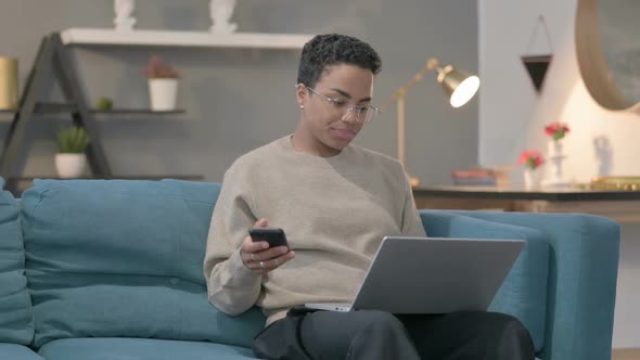 African Woman with Laptop Using Smartphone on Sofa