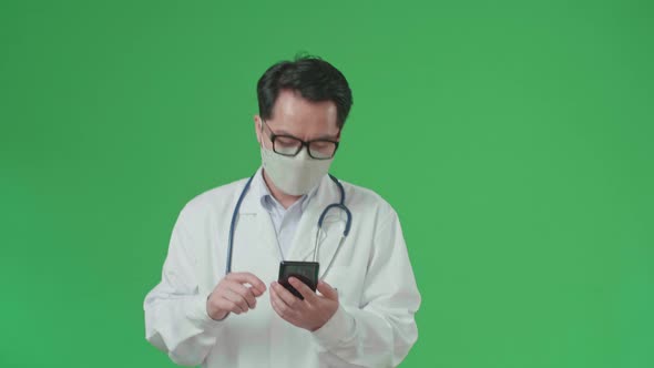 Asian Doctor With Stethoscope Wearing Mask, Using Mobile Phone While Walking On Green Screen