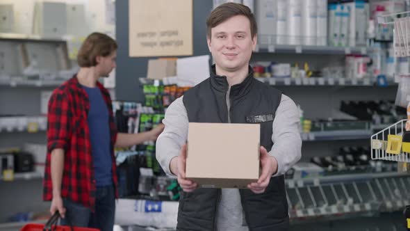 Smiling Man Stretching Box at Camera with Blurred Customer Choosing Tools at Background