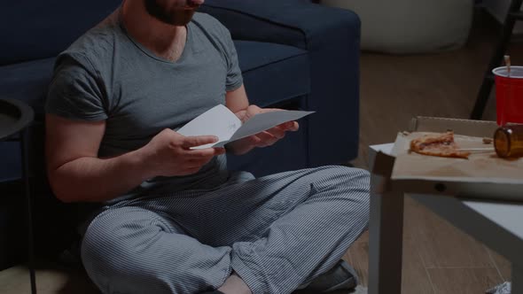 Shocked Man Sitting on Floor at Home Reading Bad News Holding Documents