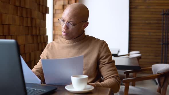 A Man with a Serious Expression Works with a Laptop and Documents in a Cafe