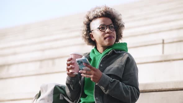 Happy adult African woman wearing casual clothes texting on phone with cup of coffee