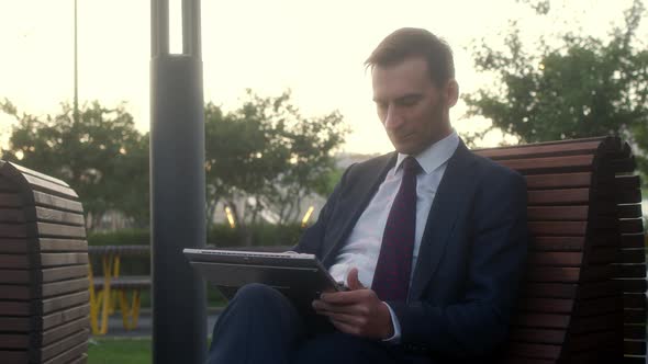 Businessman in Official Dark Blue Suit Sits on Bench in Park Before Meeting with Partners and is