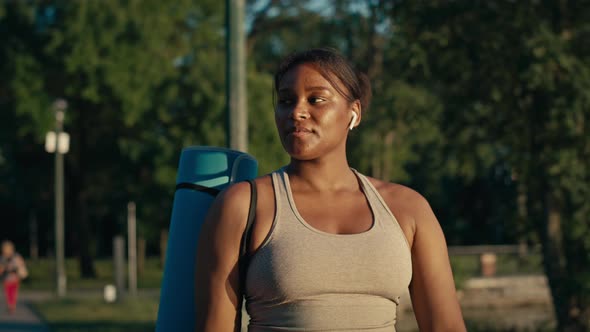 Plus size African-American woman walking with exercise mat through the park in a summer day