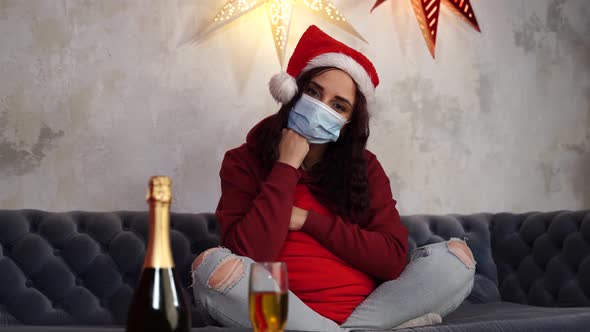 Young Woman in Medical Mask and Santa Claus Hat Sitting on Sofa in Room