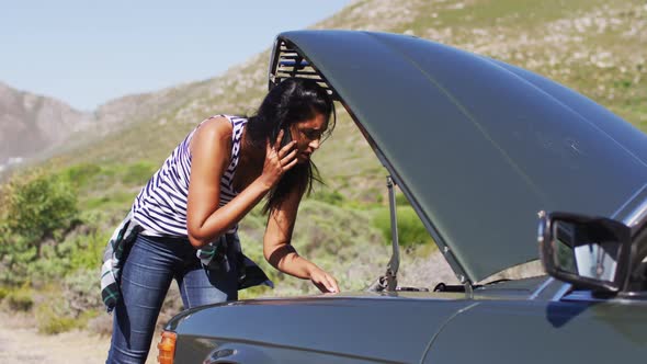 African american woman talking on smartphone while standing near her broken down car on road