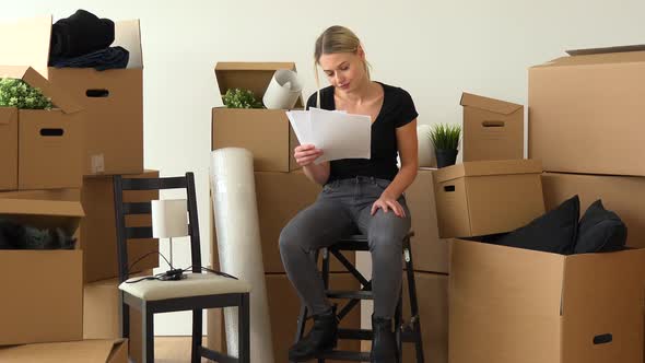 A Moving Woman Sits on a Chair in an Empty Apartment and Reads Bunch of Papers, Surrounded By Boxes