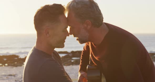 Happy caucasian gay male couple sitting on car playing guitar and kissing at the beach