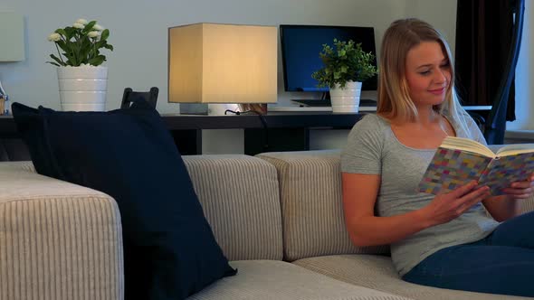 A Young, Beautiful Woman Sits on a Couch in a Living Room and Reads a Book