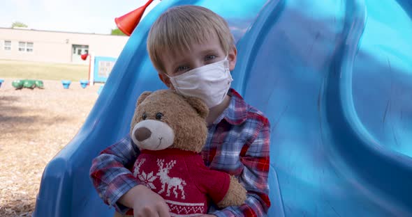 Little boy in a mask cuddling his teddy bear at a playground.