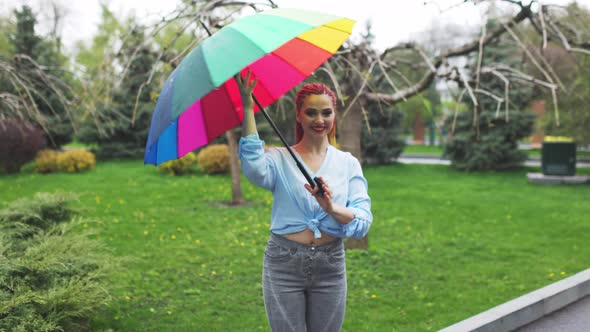 A Cute Girl with Multicolored Braids and Bright Makeup in a Bluish Shirt Posing with a Rainbow