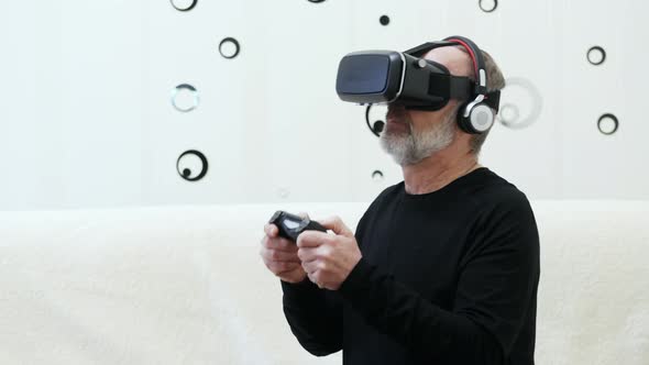 Middle-aged Man with Grey Beard Play in Virtual Reality Video Game with Wireless Headphones, Gamepad