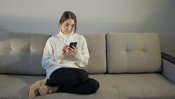 Pretty Girl in Sweater Sitting on Sofa and Read Messages on the Phone