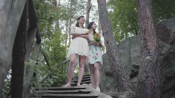 Two Young Women in Short Dresses with Flowers Standing on the Stairs and Looking at Amazing Forest