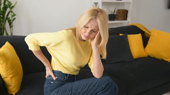 Upset Mature 50s Woman with Blondehair Suffering From Back Pain Sitting on Sofa