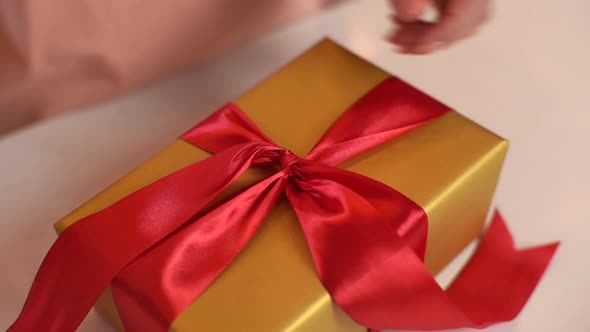 Closeup Top View Hands of Unrecognizable Young Woman Wrapping Golden Christmas Gift Box Tying Red