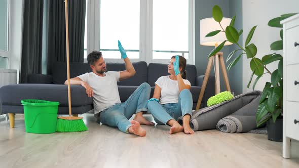 Tired Couple Sits on the Floor in a Room and High Five Each Other After They Finish Cleaning the