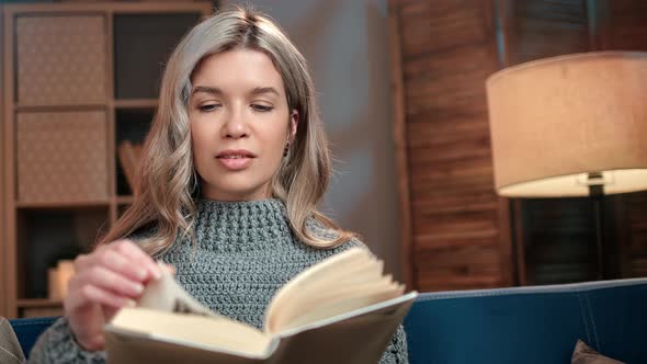 Smiling Young Blonde Female Enjoying Reading Interesting Book Turning Paper Page at Cosiness Home