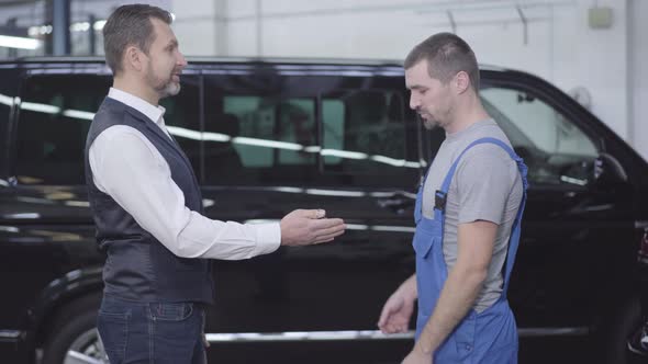 Side View of Caucasian Man Giving Keys From His Car To Auto Mechanic. Adult Businessman Talking with