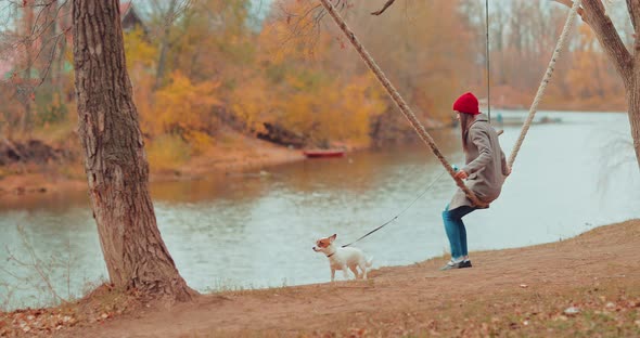 Girl is Sitting on a Swing By the River and Swinging on a Rope Holding a Dog on a Leash