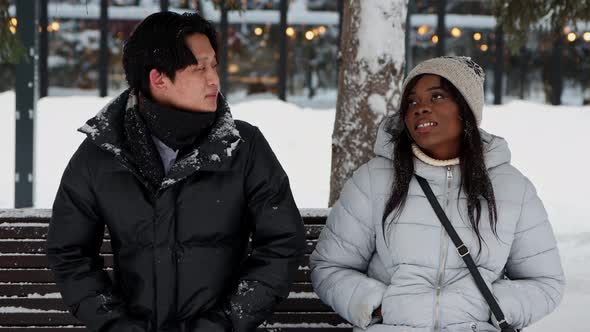 Young Asian Man Talking with His Black Girlfriend Sitting on Bench in Winter Park