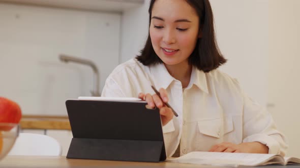 Positive Asian woman writing something and looking at the tablet