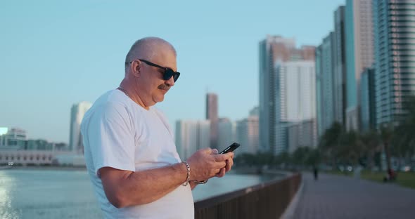 Bearded Senior Man Standing Outdoors Using Smartphone. Retired Male Reading News, Chatting