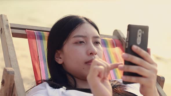 Asian woman feeling relax lying on the deck chair on the beach and using smartphones.