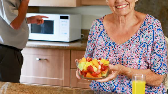 Senior woman holding bowl of fruit while man talking on mobile phone in kitchen