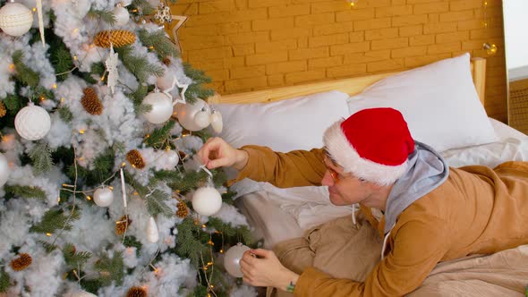 Man in Santa Hat and Glasses Lying on Bed and Decorating Christmas Tree