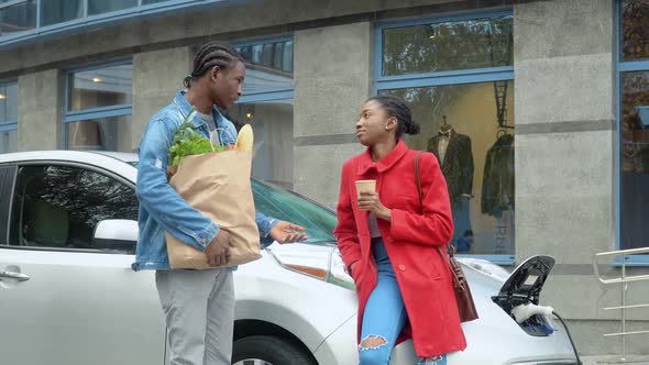 African American Couple Stands with Groceries Near Electric Car. Charging Electric Car