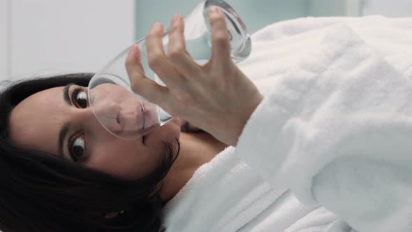 Vertical video of thirsty smiling 35s woman in white bathrobe drinking water