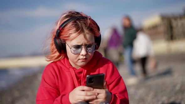 Stylish Teen Girl with Glasses Listens to Music with Headphones Uses a Smartphone on a Walk By the