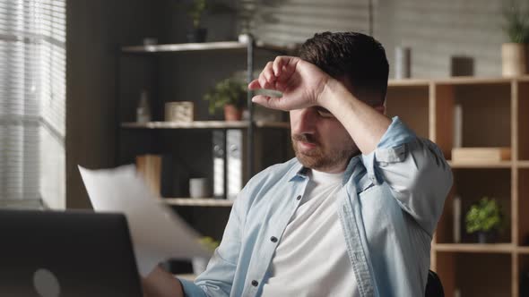 Unhappy Young Man Sitting in Hot Workplace and Using Paper for Fan