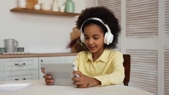 Little African American Girl in Big White Headphones Plays a Game Using a Digital Tablet