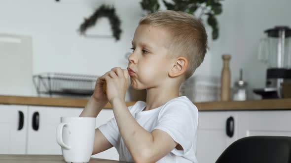 A Blonde Boy Child Eats Lunch in A Bright Kitchen, Eats A Pie And Drinks Tea or Coffee or Cocoa