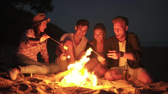 Young Cheerful Friends Sitting By the Fire on the Beach in the Evening Cooking Marshmallow on Sticks