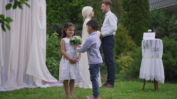 Cute Middle Eastern Boy Giving Wedding Bouquet to Pretty Girl on Marriage Ceremony Outdoors