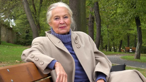 An Elderly Woman Sits on a Bench in a Park and Smiles at the Camera