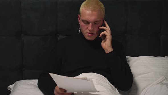 Handsome Homosexual Man Working with Documents on Bed Talking on Phone Before Going To Sleep at Home